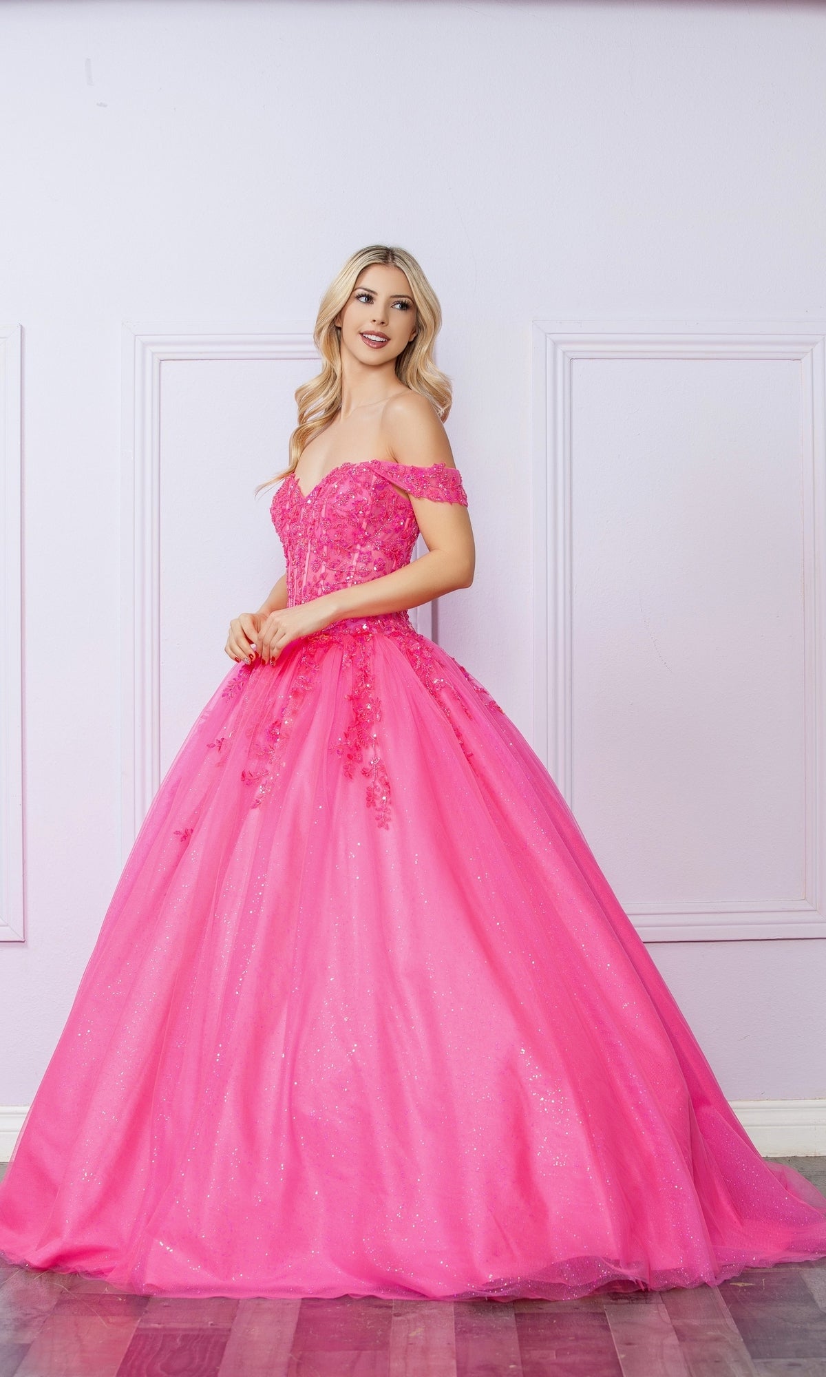 A woman in the Bright Off-the-Shoulder Long Prom Ball Gown H1349 stands against a light purple wall with white trim, smiling and gazing to the side.